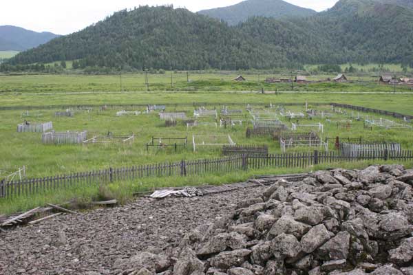 A small fenced cemetery with stones on a kurgan in the foreground and mountains in the background