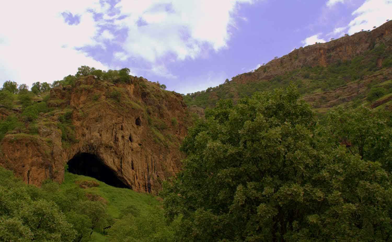 A cave opening in a rocky hill with green trees visible and a blue cloudy sky