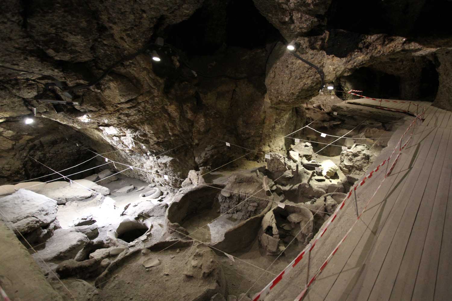 An archaeological excavation area with exposed storage pits inside of a cave, with a boardwalk flanking it at right.