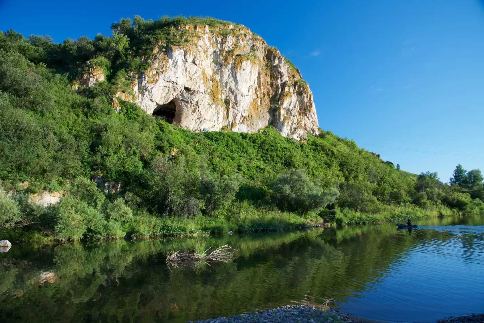 Chagyrskaya Cave in a sheer limestone cliff overlooking a river with blue sky