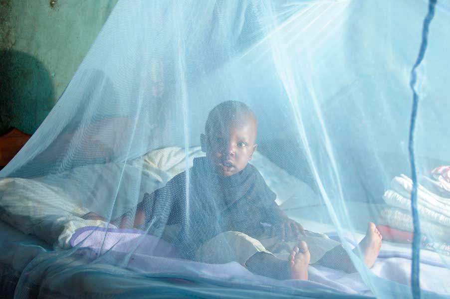 A young child sits looking at the camera on a bed beneath a mosquito net