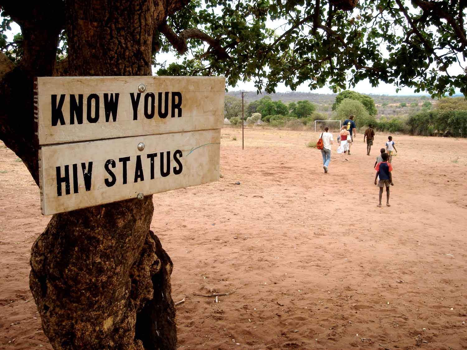 Sign nailed to a tree that reads "Know your HIV status" on a dirt soccer field with kids playing in background.