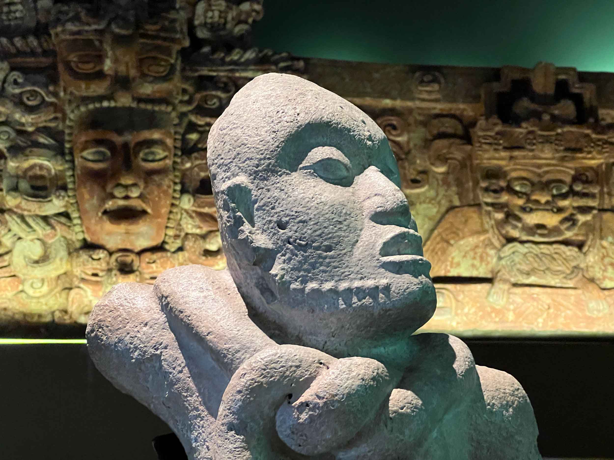 Head and shoulders of a light stone bust with a red and green colored frieze in the background, with faces and elaborate carving