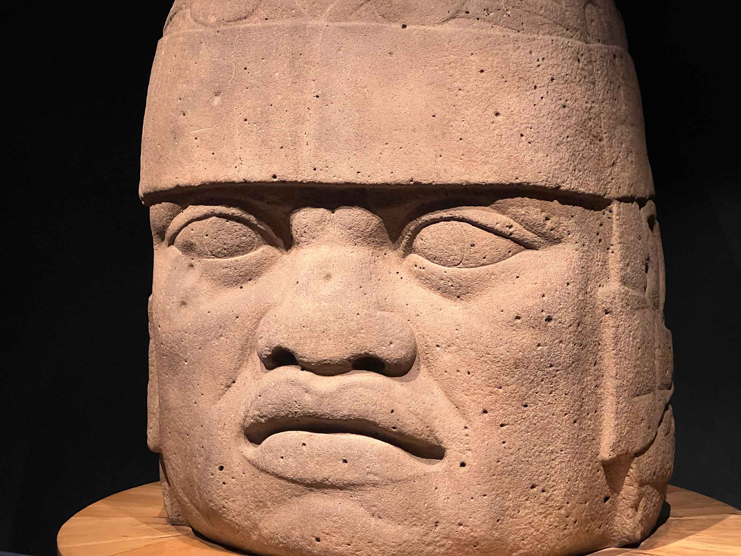 Very large head with headdress carved from stone, sitting upon a round platform