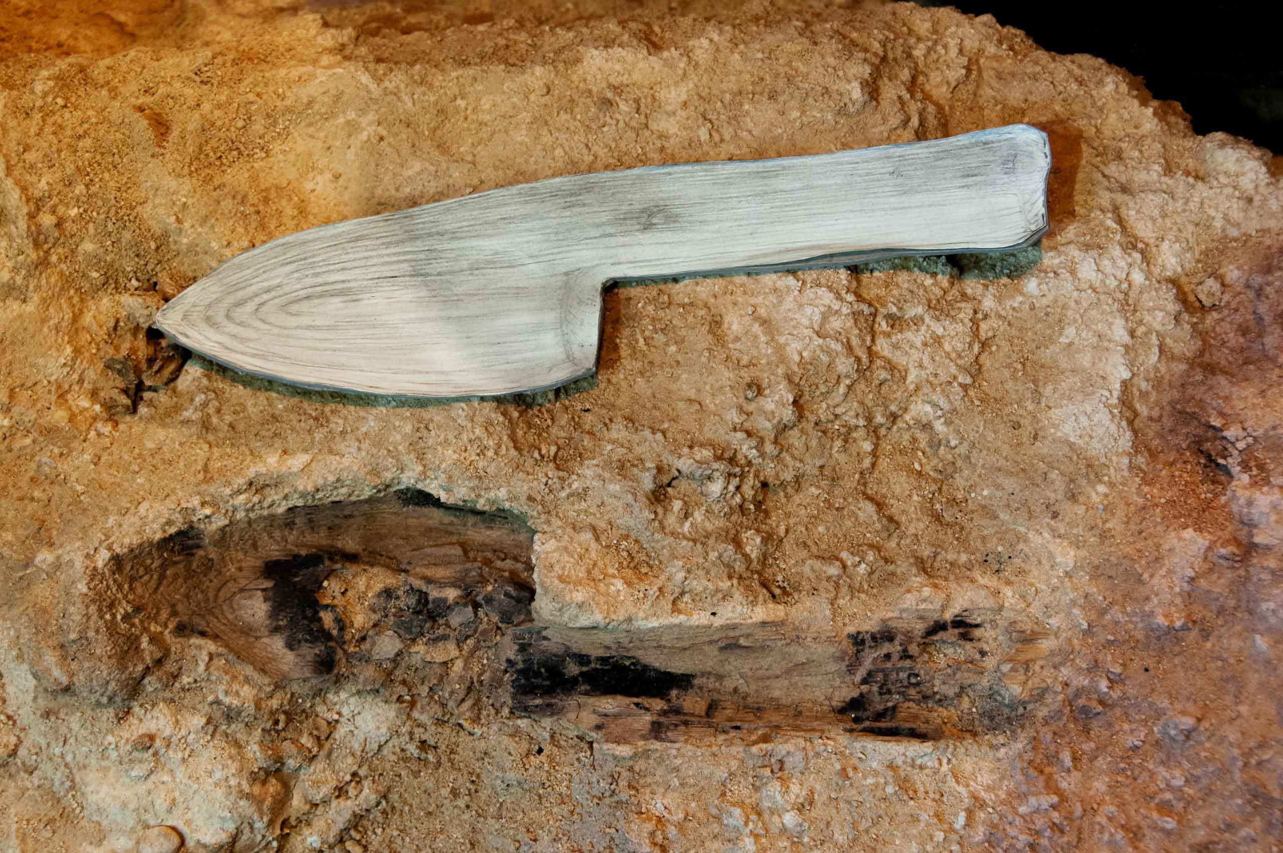 An imprint within an archaeological deposit in the shape of a knife or spade, with a wooden object of the same shape pictured above it