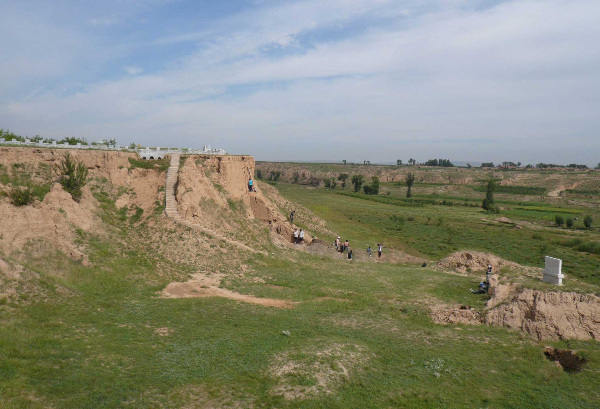A sharp cliff face composed of tan sediment, with archaeologists working on a dig and surrounding green grassy valley