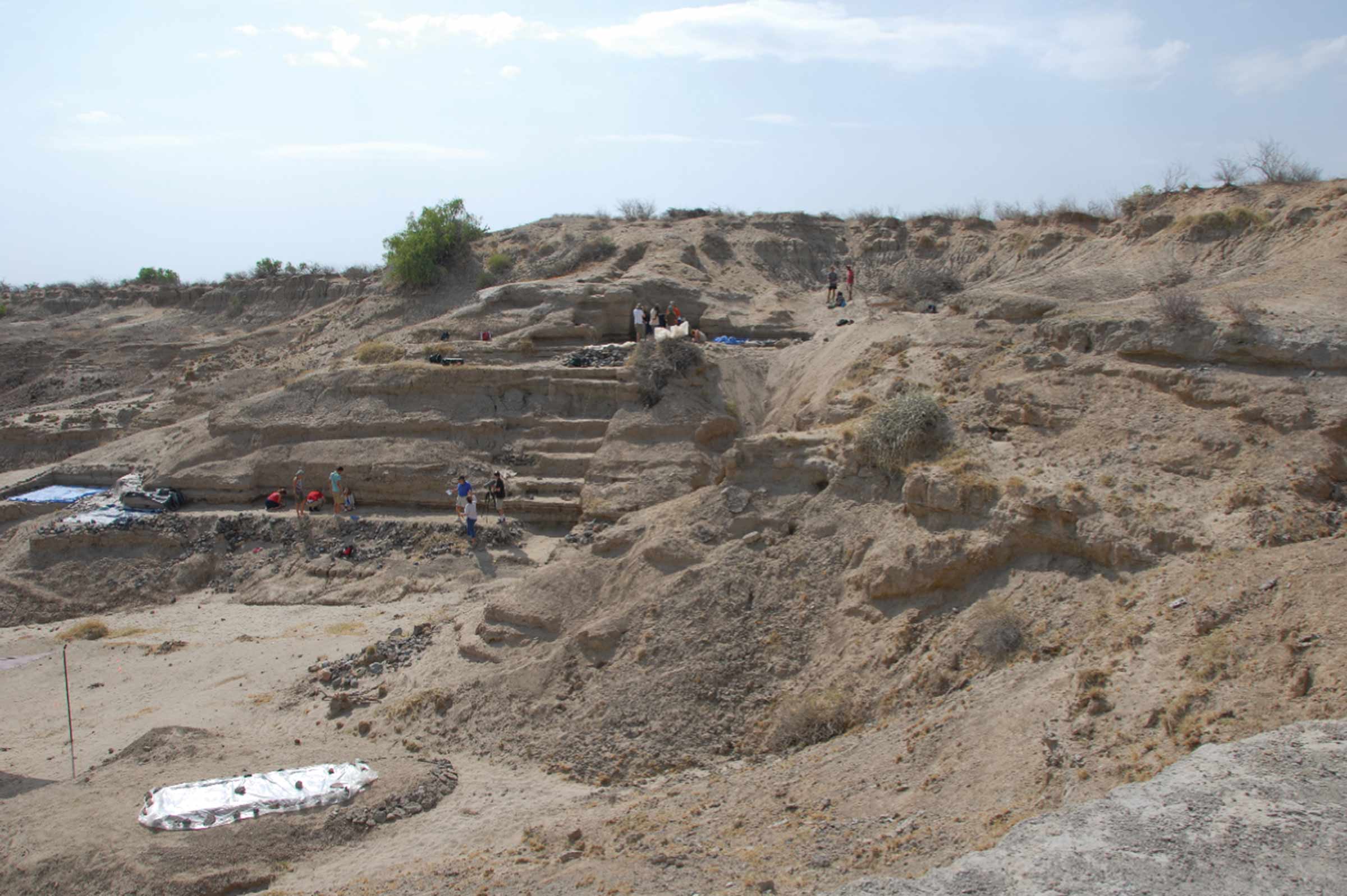 Tan hills with little vegetation, one heavily altered by digging a series of flat surfaces along its slope. Archaeologists stand on some of these surfaces.