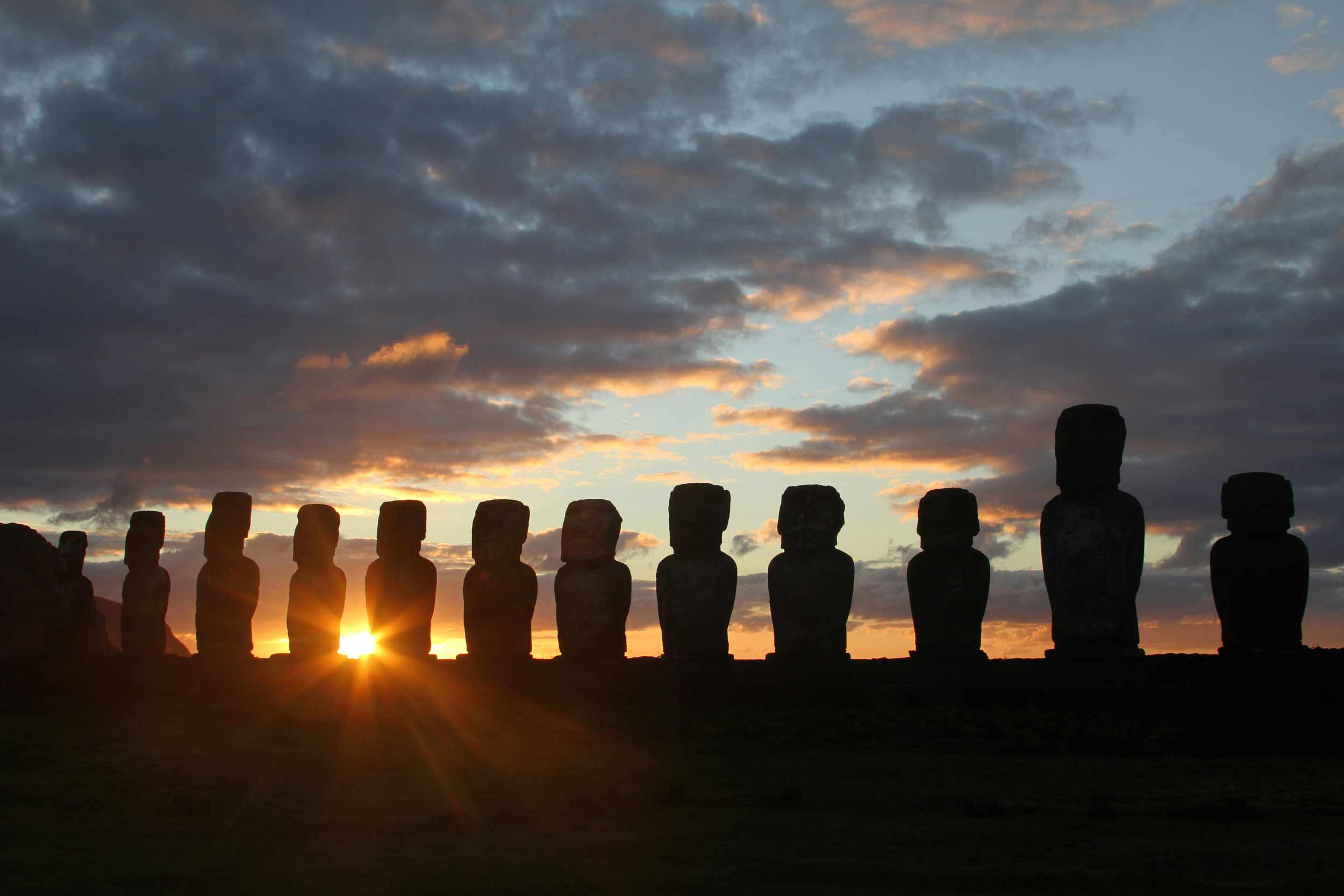 A line of enormous human statues, silhouetted by the sunset behind them.